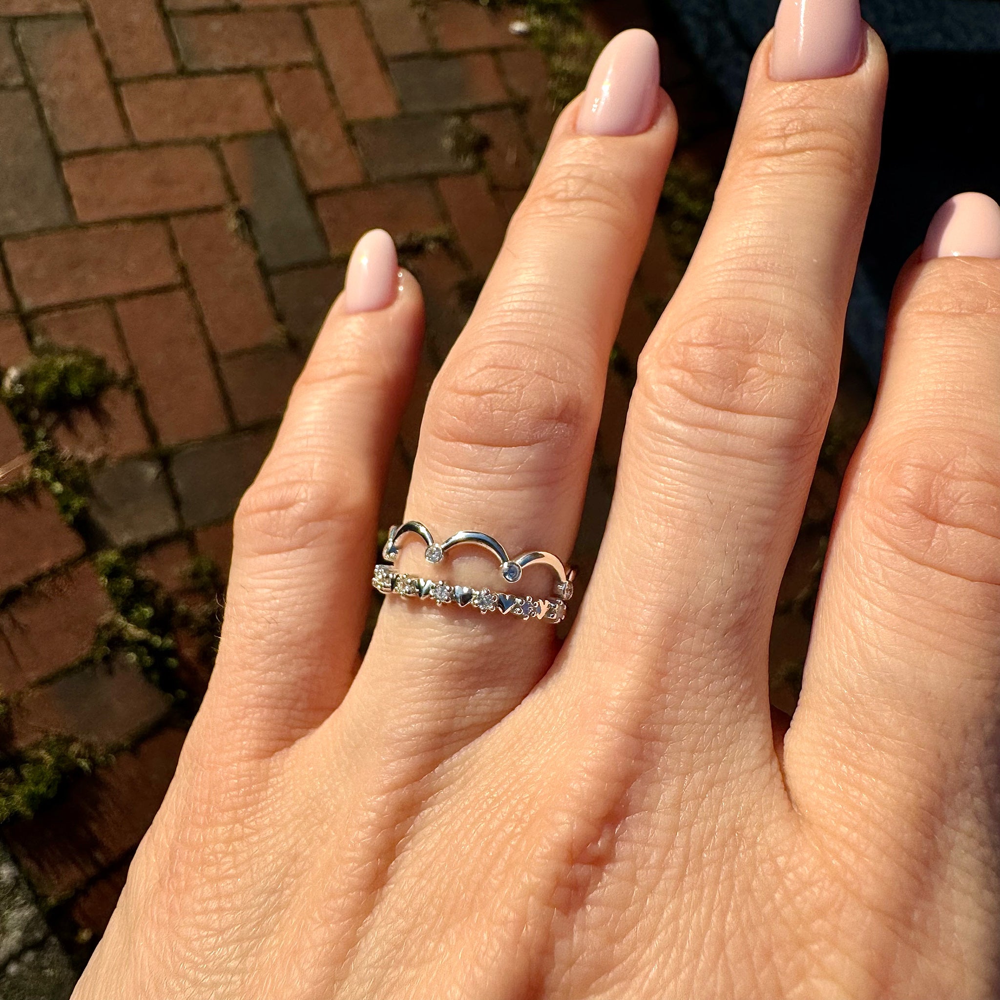 A detailed close-up photograph of a woman's neatly manicured hand with glossy light nude nail polish. Her ring finger is adorned with an elegant stack of two gold bands. The top band is a unique 14K gold eternity ring featuring an intricate alternating pattern of small lab-grown diamonds and high-polish solid gold heart shapes (the 