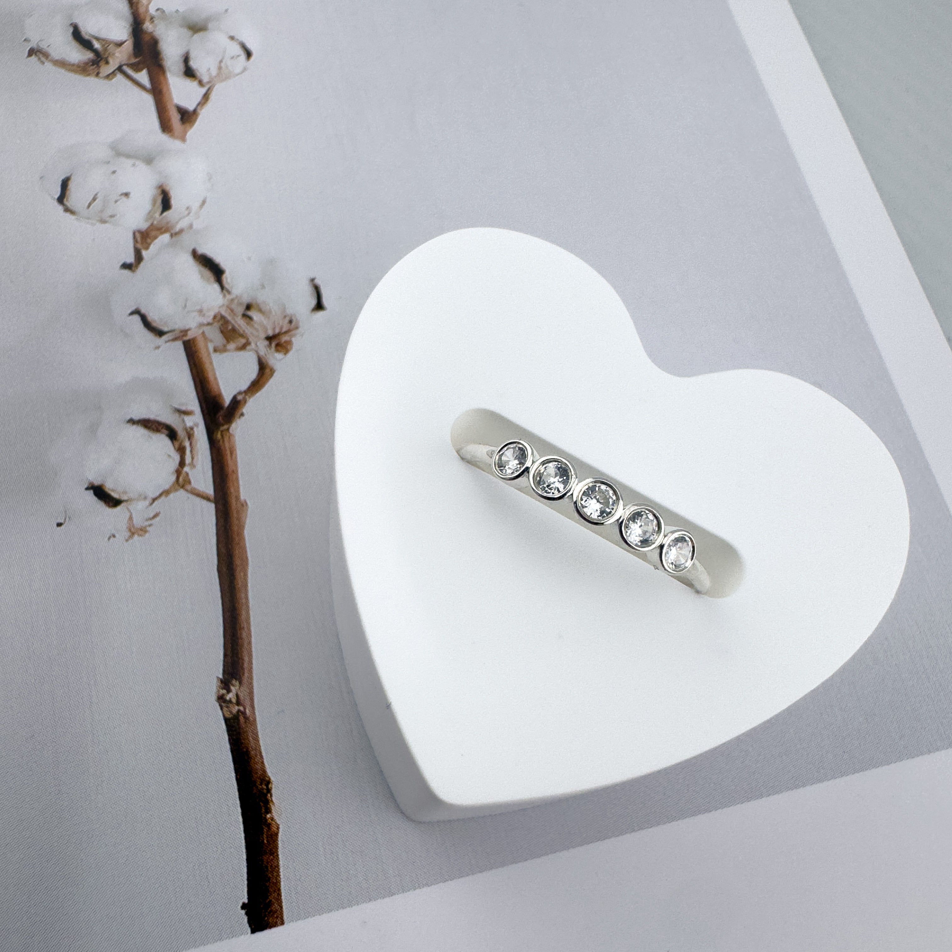 A professional product photograph of the Luna 14K white gold stackable ring with five bezel-set lab-grown diamonds, elegantly displayed on a white heart-shaped stand next to a cotton branch.